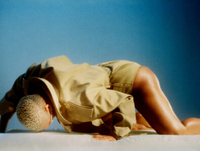 A person with short, light-colored hair is positioned in an elegant, dynamic pose on a neutral surface against a gradient blue background. They wear a stylish, tan jacket, with the fabric gently folding and catching ambient light. The scene exudes a sense of movement and contemporary fashion, highlighted by the shadows and soft, contrasting colors.