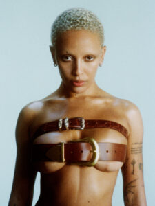 A person with short, curly hair poses confidently. Their torso is adorned with two wide, brown leather belts positioned across the chest. Each belt features a prominent buckle, adding a bold and unconventional fashion statement. The background is a muted shade of blue, highlighting their tattoos on the arms and emphasizing the creative styling.