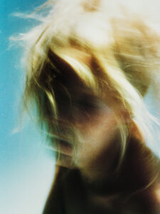 A blurred, dreamy portrayal of a person with light hair swirling around, creating a sense of motion and ethereal beauty. The soft, warm tones intertwine with a gentle blue background, enhancing the abstract, artistic feel. The image captures a fleeting moment, evoking emotions of freedom and fluidity, inviting viewers to interpret its essence personally.