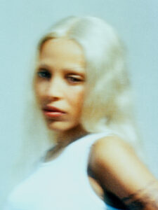 A person with long, light-colored hair stands against a soft blue backdrop, their figure artfully blurred to create an ethereal effect. They are wearing a white sleeveless top, slightly turned to the side, gazing enigmatically past the viewer. The image combines a dreamy ambiance with warm, gentle lighting, enhancing the overall sense of mystery and allure.