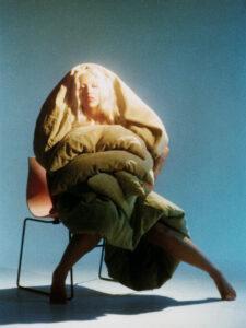 A person with long blonde hair is seated on a simple chair. They are draped in a thick, wrinkled green fabric that envelops their upper body, creating a cocoon-like appearance. The background is a gradient of blue to green hues, casting a tranquil, dreamy atmosphere. The lighting is soft, highlighting the serene expression on their face.