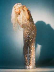 A person with long, flowing blonde hair stands gracefully, captured mid-movement with a slight bend forward. They wear a shimmering, translucent outfit that glimmers with reflective elements, catching the light beautifully. The background features a soft gradient of cool tones, enhancing the ethereal quality. Their expression is serene, adding to the overall dreamy, otherworldly atmosphere.