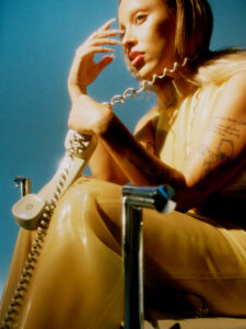 A person with long hair and red lipstick sits in a chair, holding a vintage phone with a coiled cord. They wear a sleeveless, glossy beige outfit, showcasing tattoos on their arms. Their left hand rests on their chin, creating a thoughtful pose. Warm lighting casts shadows, enhancing the overall retro atmosphere against a blue background.