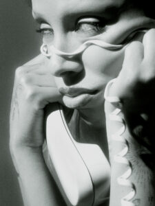 A close-up monochromatic composition shows a person holding a vintage telephone receiver to their face. A telephone cord is wrapped around their eyes, casting intricate shadows. Their expression seems contemplative, with tattooed arms resting on the face. The light accentuates the contrast between smooth skin and textured shading, creating an artistic, surreal atmosphere.