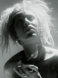 A black-and-white composition showcases a figure with messy, light hair against a stark backdrop. The focus is soft, with a pronounced play of light and shadows, accentuating pensive expression. Hand movements are visible with smoke curling upwards, creating an ethereal atmosphere. The overall mood is contemplative and slightly mysterious, capturing a moment of introspection.