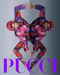 A mirrored figure forms a heart like silhouette through carefully posed arms and legs. The person wears a vibrant, padded hooded jacket and patterned leggings in swirling purples, reds, and greens. Their gaze is direct, makeup softly defined. The symmetrical composition feels graphic, playful, and sculptural, blending fashion with body geometry, rhythm, balance, bold colour, contemporary fantasy.