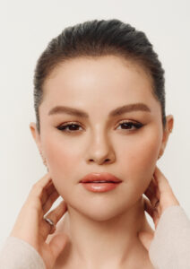 A person with smooth skin and subtle makeup is depicted. The hair is dark and neatly pulled back, accentuating features like arched eyebrows and glossy lips. The gentle expression is emphasized by softly blushed cheeks. Hands delicately frame the face, one wearing a ring, with a neutral background adding to the serene and elegant atmosphere.