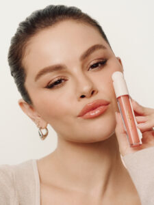A woman with dark hair pulled back gazes forward, showcasing a natural makeup look with defined brows and glossy lips. She holds a tube of lip gloss close to her face. Wearing a light-colored top, she exudes a serene and confident expression. Her earrings add a touch of elegance to her appearance.