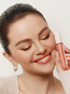 A person with glowing skin and styled dark hair smiles gently with eyes closed. They hold a tube of pink lip gloss near their face. The makeup is natural, featuring peachy tones on the lips and lightly blushed cheeks. They wear small hoop earrings and a soft, neutral-colored top, creating a harmonious and elegant look.