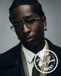A close-up portrait shows a man with neatly braided hair, wearing slim rectangular glasses and a single hoop earring. His expression is calm and introspective, gaze slightly angled. He is dressed in layered tailoring, including a collared shirt, tie, and structured outerwear. Soft, controlled lighting highlights skin texture and contours, creating a refined, moody atmosphere against a neutral background.
