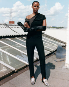 A person stands confidently on a rooftop, dressed in an asymmetrical black outfit with long gloves. The outfit has a single sleeve and is paired with metallic silver shoes. In the background, a cityscape with clouds and a transmission tower is visible, creating a juxtaposition between the urban environment and the stylish, futuristic attire.