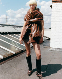 A person with short blonde hair stands confidently on a rooftop. They are wearing a unique, oversized brown puffer top and shorts, paired with tall black boots featuring decorative chains. The sky is filled with fluffy white clouds, and buildings with corrugated rooftops and a brick structure are visible in the background, adding an urban contrast.