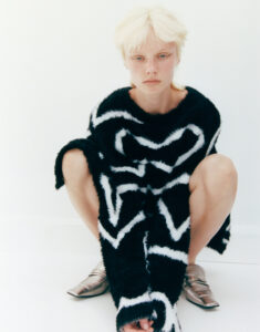 A person with short, light hair sits on a plain background. They are dressed in a bold, fuzzy black-and-white patterned garment that covers their arms and torso. Their feet are bare aside from metallic shoes. The expression is calm and introspective, and the overall composition highlights the texture and contrast of the attire against the light setting.