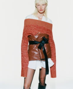 A person with light, shoulder-length hair wears a unique outfit combining a red, off-the-shoulder sweater with long sleeves, a brown leather waist jacket, and a black ribbon belt. Underneath, a white garment subtly peeks out. The overall look is edgy and fashion-forward, completed by knee-high black boots and a neutral expression. The background is plain white.