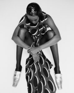 A black-and-white portrayal features a person sitting with legs crossed and arms resting on knees. The dress has a bold, circular pattern, and the footwear is shiny with a square toe design. The individual looks downward, creating a contemplative mood. The overall aesthetic is modern and minimalist, contrasting elegant attire with a simple, clean background.