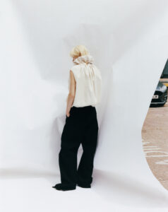 A person stands with their back turned, wearing a sleeveless, high-collared white top and loose black pants. Their blonde hair contrasts with the soft folds of the white fabric they are facing. The setting includes a draped white backdrop, partially revealing a sliver of urban pavement and a parked car in the background, adding depth.