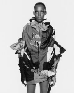 A person stands, centered in a monochrome setting, wearing an oversized, patchwork-style garment with various textures and patterns. The garment appears to be a mix of different materials, featuring layers and exaggerated proportions. The person's expression is composed and direct, with a minimalist background that enhances the focus on the intricate details of the outfit.