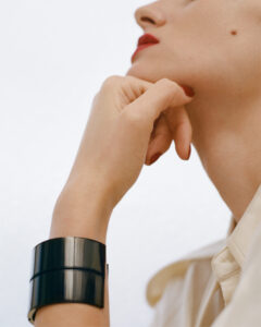 A person poses elegantly, resting their chin on their hand. The focus is on the side profile, with vibrant red lips and a small mole visible. They wear a wide, glossy black bracelet and have matching red nail polish. The background is softly blurred, emphasizing the cream-colored fabric of their attire and the refined aesthetic.