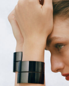 A close-up captures an individual with light skin, showcasing their right profile. Their hand rests on their forehead, with short hair brushed back. They wear two sleek black bracelets on the wrist. The person's lips are painted a bold red, and their green eye is prominently observed. The background remains a soft, neutral tone.