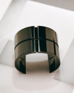 A sleek metallic cuff bracelet is positioned against a smooth surface. The reflective black finish features two parallel grooves encircling the band. Its open-ended design forms a graceful arc, casting subtle shadows upon the pale background. The piece exudes contemporary elegance, exemplifying minimalist style, and echoes modern architectural influences through its clean lines and polished exterior.