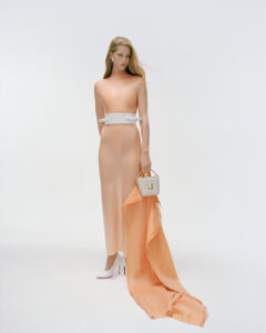 A person stands elegantly, wearing a long, peach-colored dress with a dramatic train flowing on the ground. The outfit is complemented by a white belt and matching high-heeled shoes. They hold a small, structured handbag with a short handle. The background is minimalist and clean, focusing the viewer's attention on the stylish ensemble and poised stance.