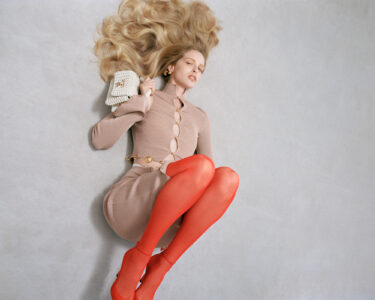 A woman lies on a light surface with flowing blonde hair spread around her. She wears a beige dress with large buttons and vibrant red tights, coupled with matching red heels. Her eyes are closed, holding a small, vintage-style handheld mirror near her hip. The scene conveys elegance, with a minimalist and fashion-forward aesthetic.