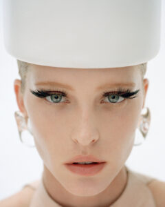 A person gazes directly ahead, showcasing bold eyelashes and clear, light-colored eyes. They wear large, circular earrings and have neutral-toned lips. Their hair is tucked under a cylindrical white headpiece, creating a distinctive, modern look. The overall composition emphasizes minimalism and strong facial features, highlighting an avant-garde fashion aesthetic with a focus on elegance and simplicity.