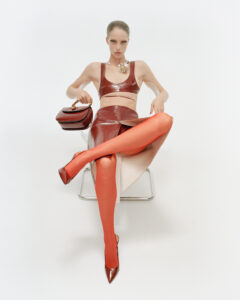 A person with a slicked-back hairstyle lounges on a modern chair, exuding confidence. They're dressed in a shiny, dark red ensemble, consisting of a crop top and skirt. Bright orange stockings and pointed heels add bold flair. Accessories include a necklace and a handbag perched on one arm, completing the strikingly fashionable appearance.