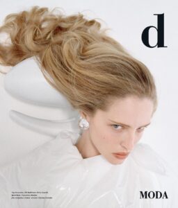 A person with long, flowing blonde hair poses against a light background. Their expression is serious, with piercing blue eyes gazing intently forward. They wear a large, clear earring and are draped in a shiny, white material, giving a modern, avant-garde vibe. The setup highlights a blend of fashion and art with minimalistic elegance.