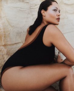 A person with light skin is posed against a textured stone backdrop. They have medium-length, wet hair slicked back. The individual is wearing a black one-piece garment with a low-cut back. Their make-up is minimal yet striking, with notable eye shadow. The pose is dynamic, emphasizing curves and angles, creating a powerful and confident aesthetic.