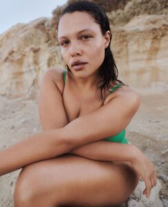 A person with dark hair and freckles gazes directly, seated with arms crossed over bare knees. They wear a green outfit, sitting against a rocky, beige cliff. The calm, earthy tones of the background contrast with their contemplative expression. The scene conveys a sense of natural beauty and serenity, merging the human presence with the rugged landscape.