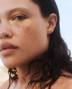 A person with wet, dark hair partially covering their shoulder is seen closely. They have warm-toned skin with subtle freckles and wear shiny gold eyeshadow that highlights their eyes. The expression is serene and contemplative, and the soft natural lighting enhances the golden and warm hues of their makeup and skin, suggesting a calm and relaxed atmosphere.