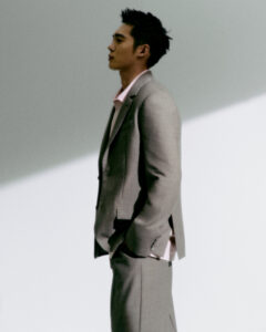 A person stands in profile against a minimalist backdrop with angled lighting. The individual is dressed in a tailored light gray suit, paired with a pale pink shirt. Their hands are casually placed in the suit pockets. The setting features a blend of shadows and highlights, creating a sophisticated and contemporary atmosphere. Hair is styled upward neatly.