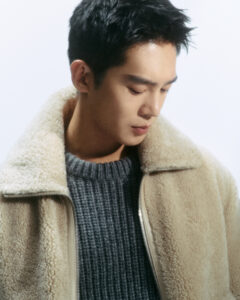 A young person with short dark hair looks down thoughtfully. They wear a cozy beige fleece jacket over a textured gray sweater. The lighting is soft and ambient, emphasizing the textures of the clothing while casting gentle shadows on the person's face. The overall mood is calm and introspective, capturing a moment of quiet reflection.