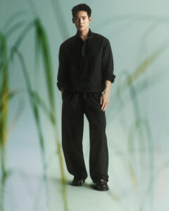 A person stands with hands in pockets, dressed in a relaxed black outfit consisting of a loose-fitting long-sleeve top and matching pants. The background is softly blurred, featuring green, plant-like shapes. The overall atmosphere is calm and minimalistic, with a muted color palette that creates a serene, natural ambiance. The composition feels balanced and visually soothing.