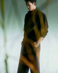 A young man with short dark hair stands contemplatively, wearing a dark, loose-fitting outfit. His left hand rests casually in his pocket, while dried plants blur the foreground. The background features a soft gradient of muted green to pale hues, creating a tranquil and introspective atmosphere. Soft lighting enhances the serene and sophisticated mood of the scene.