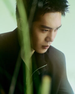 A thoughtful man gazes downward with a contemplative expression, partially obscured by green foliage in the foreground. He wears a dark jacket with a zipper, and the tone set by the soft lighting creates a tranquil, introspective atmosphere. The overall composition conveys a sense of calm reflection, enhanced by the blurred greenery around him.