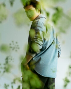 A person with a short haircut is wearing a light denim jacket and dark pants. Their expression is thoughtful, as they stand among blurred green foliage. The overall atmosphere is serene and natural, with soft lighting that highlights the gentle contours of their face. The composition blends fashion with elements of nature, creating a harmonious scene.