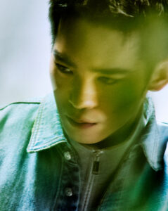 A young individual is captured in a thoughtful moment, partially obscured by soft, green foliage. Dressed in a light denim jacket with a gray shirt underneath, their expression seems introspective. The lighting is subdued with a gentle, natural glow, creating a serene and contemplative atmosphere around the subject.