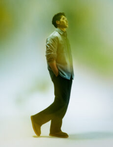 A person stands confidently with a relaxed posture, hands in pockets, slightly tilting their head back, and appears deep in thought. They wear a casual outfit consisting of a light jacket and dark pants. The overall scene has a soft, dreamy quality, with a green filter adding a serene, contemplative mood to the setting.