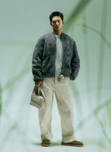 A person stands confidently, wearing a quilted jacket and light-colored pants. They hold a small bag in one hand, offering a sense of style and casual elegance. The background is soft and blurred with hints of green, adding a calming atmosphere. The footwear is casual, complementing the overall relaxed and modern look.