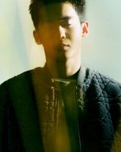 A young man stands with his eyes closed, partially bathed in warm, golden light. He wears a textured, dark quilted jacket over a light shirt. The composition features an interplay of light and shadow, creating a serene and contemplative atmosphere. The soft focus and muted color palette lend an introspective and almost dreamy quality to the scene.