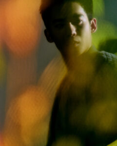A young man stands partially in shadow, his face softly illuminated, surrounded by an abstract play of lights in warm hues of yellow, green, and orange. The composition creates a mysterious and introspective atmosphere, with blurred elements adding depth and texture. Subtle contrasts between light and dark highlight the contemplative expression on his face.