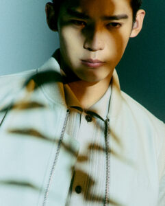 A person stands in partial shadow, with soft light casting diagonal lines across the face and upper body. The individual wears a light-colored jacket with a zip front and a collared shirt underneath. The expression is contemplative, and the overall color scheme features muted tones with a greenish tint, suggesting a calm, introspective atmosphere.