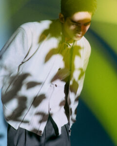 A young man leans forward, wearing a light-colored jacket over a textured shirt and dark pants. Dappled light creates intriguing shadows across his clothing and face. The background features abstract, soft-focus colors of green and blue, adding a dreamy, artistic ambiance. The composition emphasizes a moody and contemplative atmosphere, merging fashion with artistic photography.