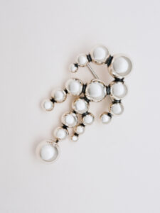 A cluster of shiny, reflective spheres of varying sizes forms a cascading design. The spheres appear metallic, with some larger and others smaller, creating a visually dynamic arrangement. The piece is elegant, suggesting a decorative or ornamental purpose, with a polished, sophisticated appearance against a neutral background. The overall composition is delicate and visually appealing.