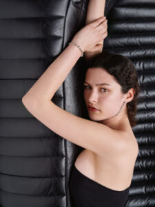 A person with curly dark hair poses in a strapless black outfit against a quilted black leather background. The person extends their arms above their head, crossing them at the wrists, adorned with a simple bracelet. The composition highlights a serene expression and sophisticated elegance, blending harmoniously with the textured surroundings and understated style.