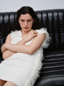 A woman with curly hair sits on a black leather sofa. She is wearing a sleeveless white dress with textured edges and has a thoughtful expression, her arms folded across her front. She accessorizes with gold hoop earrings and rings. The setting suggests a minimalist, modern aesthetic, with emphasis on texture and contrast between her attire and the furnishings.