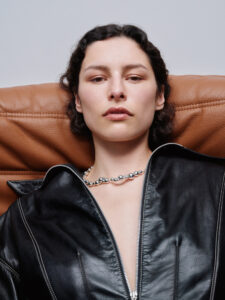 A person with short, wavy dark hair appears relaxed while leaning back against a brown, cushioned surface. They are wearing a black leather jacket with a partially open front, revealing a silver beaded necklace. The expression is calm and composed, set against a plain background that enhances the person's tranquil demeanor. The lighting is soft and neutral.