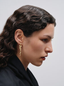 A profile view showcases a person with curly dark hair styled neatly. They wear a sleek black outfit, drawing attention to ornate gold earrings with a cascading design. The background is a simple, muted hue, emphasizing the refined elegance and contemplative expression of the individual. The composition highlights minimalistic sophistication and understated beauty.